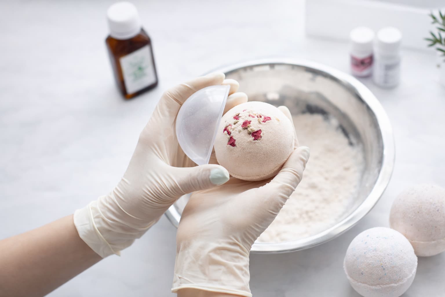 Hände mit Schutzhandschuhen halten frisch geformte Badebombe mit Rosenblüten – Badebomben selber machen und verkaufen